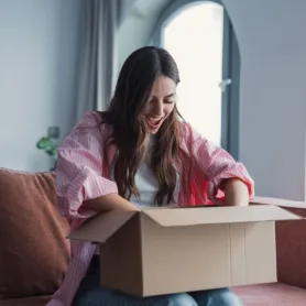 Close up overjoyed smiling young woman opening cardboard box with awaited parcel, sitting on couch at home, satisfied client received online store order, good quick delivery service concept
