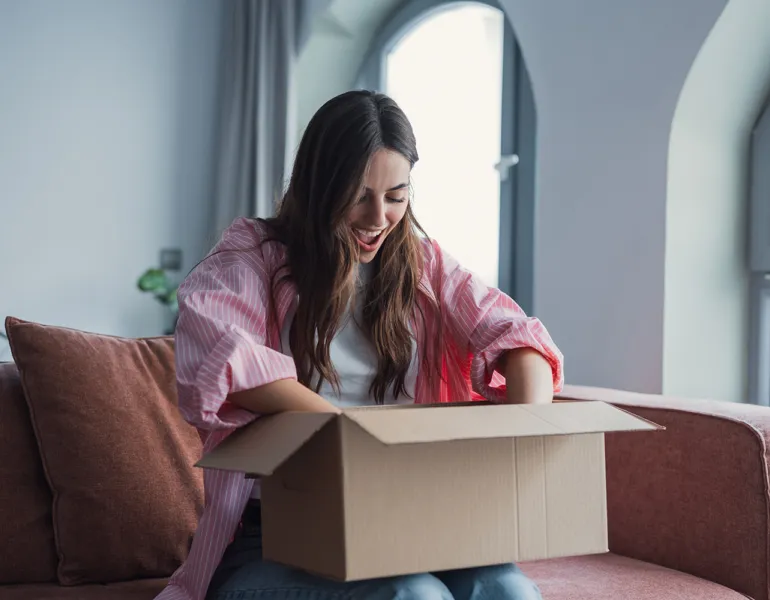 Close up overjoyed smiling young woman opening cardboard box with awaited parcel, sitting on couch at home, satisfied client received online store order, good quick delivery service concept