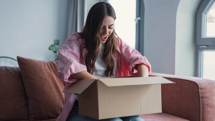 Close up overjoyed smiling young woman opening cardboard box with awaited parcel, sitting on couch at home, satisfied client received online store order, good quick delivery service concept