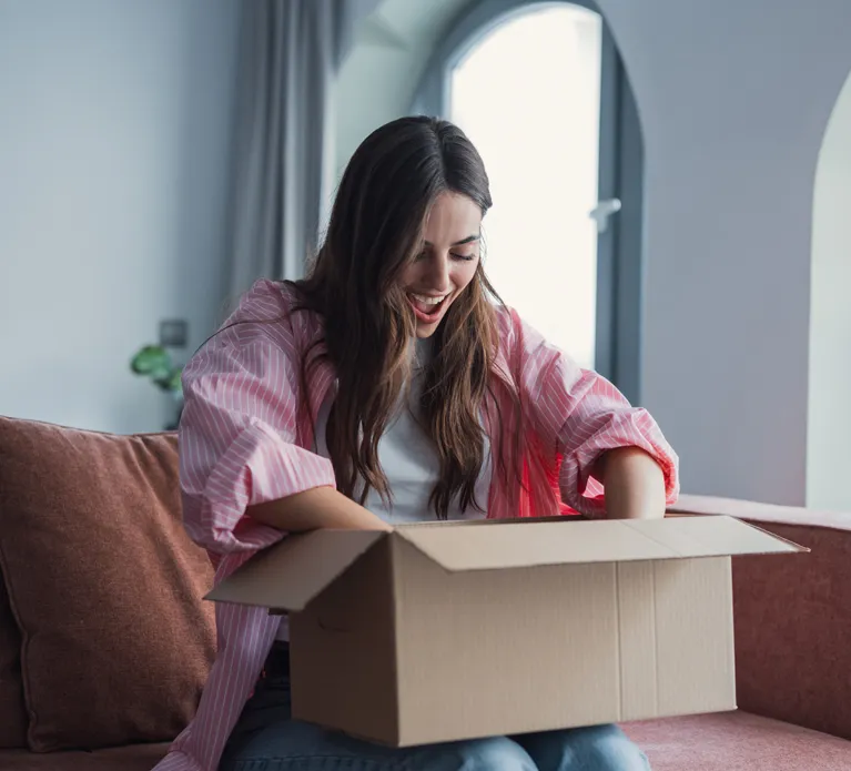 Close up overjoyed smiling young woman opening cardboard box with awaited parcel, sitting on couch at home, satisfied client received online store order, good quick delivery service concept