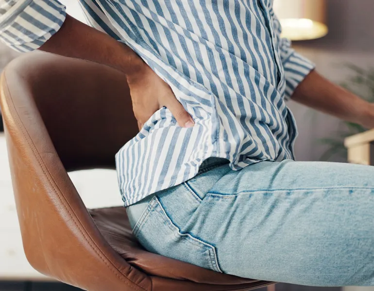 Person, hands and chair with back pain or injury for bad posture or discomfort at home office. Closeup, freelancer or remote worker with sore spine or muscle tension for inflammation or fibromyalgia