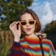 Woman, young woman, striped sweater, yellow sunglasses, smiling: early twenties female with short brown hair in colorful knit jumper and retro shades, playful surprised expression, casual shoulder