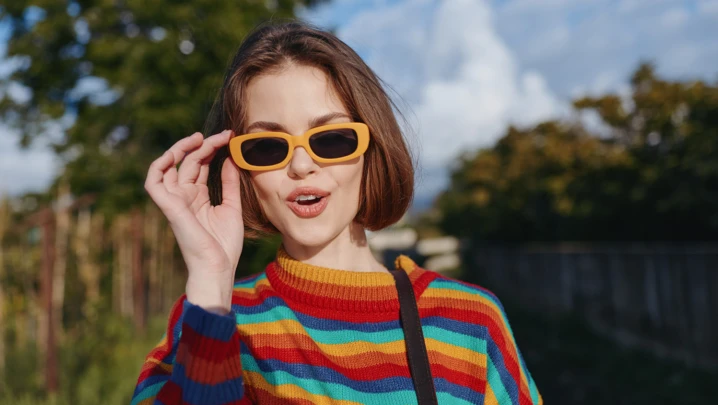 Woman, young woman, striped sweater, yellow sunglasses, smiling: early twenties female with short brown hair in colorful knit jumper and retro shades, playful surprised expression, casual shoulder