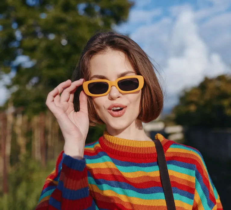 Woman, young woman, striped sweater, yellow sunglasses, smiling: early twenties female with short brown hair in colorful knit jumper and retro shades, playful surprised expression, casual shoulder
