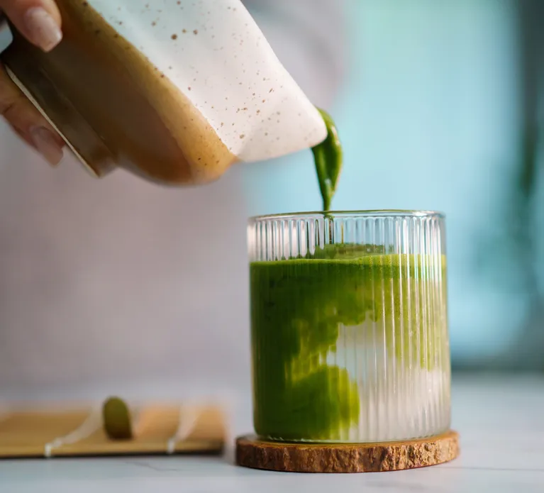 Woman pouring freshly whisked matcha into glass, mixing with coconut water. Artisan beverage preparation, Japanese tea culture, organic ingredients, and a calm home café atmosphere.