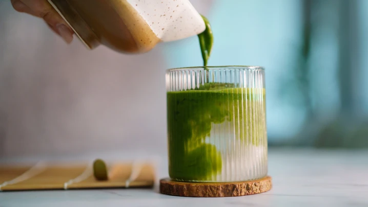 Woman pouring freshly whisked matcha into glass, mixing with coconut water. Artisan beverage preparation, Japanese tea culture, organic ingredients, and a calm home café atmosphere.