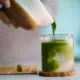 Woman pouring freshly whisked matcha into glass, mixing with coconut water. Artisan beverage preparation, Japanese tea culture, organic ingredients, and a calm home café atmosphere.