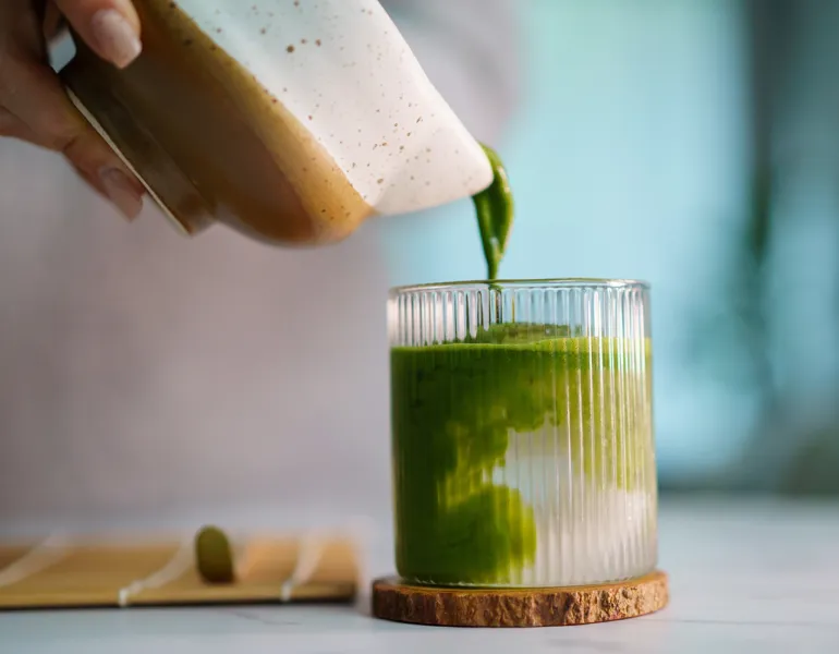 Woman pouring freshly whisked matcha into glass, mixing with coconut water. Artisan beverage preparation, Japanese tea culture, organic ingredients, and a calm home café atmosphere.